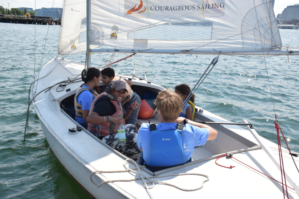 A group of people wearing life jackets, including children and an adult, are seated in a small sailboat on the water with the sail marked "Courageous Sailing.